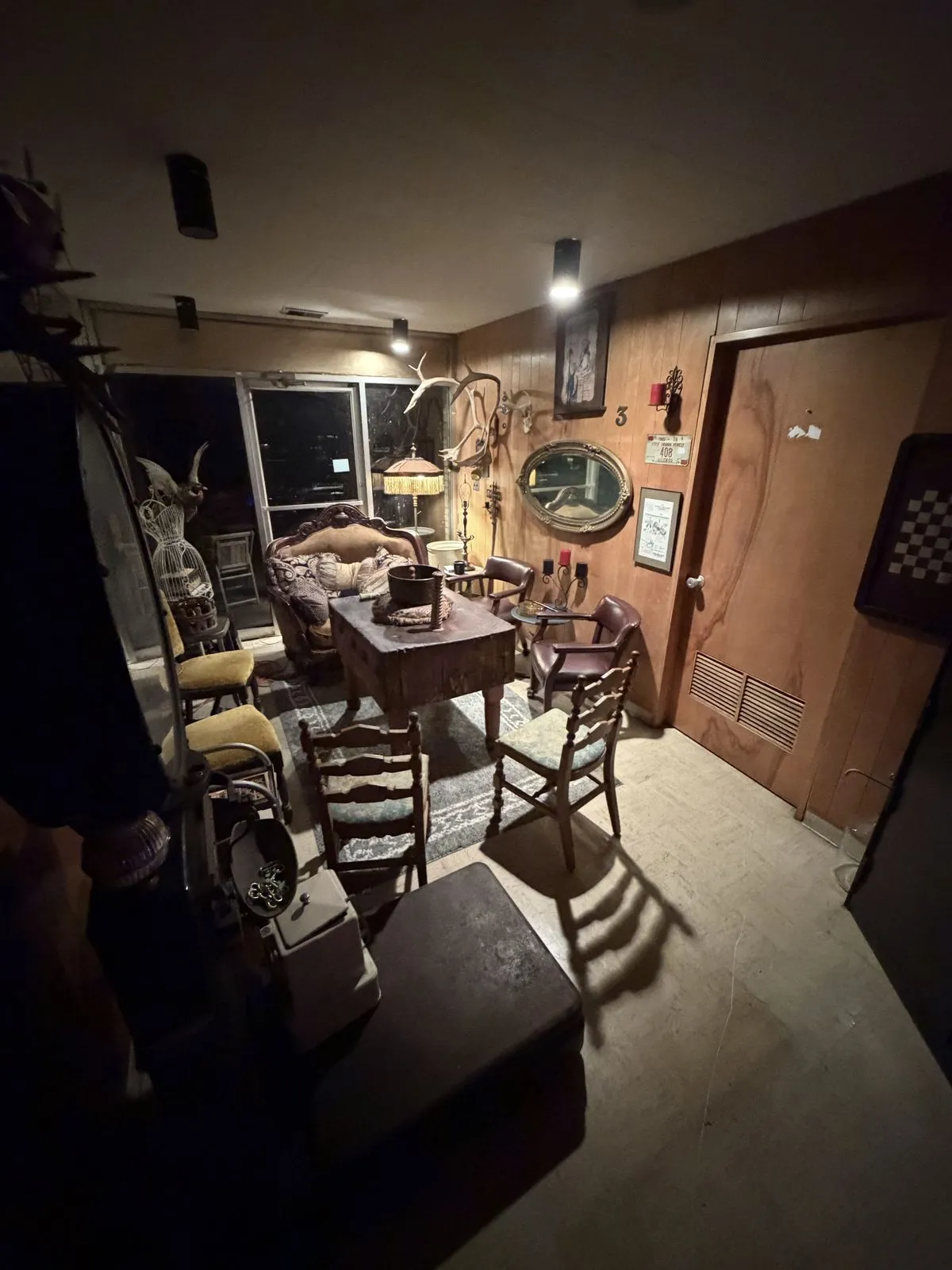 A dim parlor with a wooden table, high-backed chairs, a velvet settee, an oval mirror and antlers on the wall
